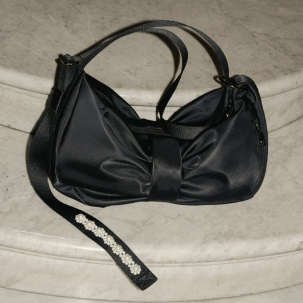 Simone Rocha Beaded Sling Bow Bag — black satin shoulder bag with structured front bow and pearl-beaded crossbody strap on marble surface