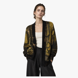 A woman modeling a black open-front cardigan with a textured, metallic gold abstract pattern, styled over a cream tank top and dark wide-leg trousers against a plain white background.