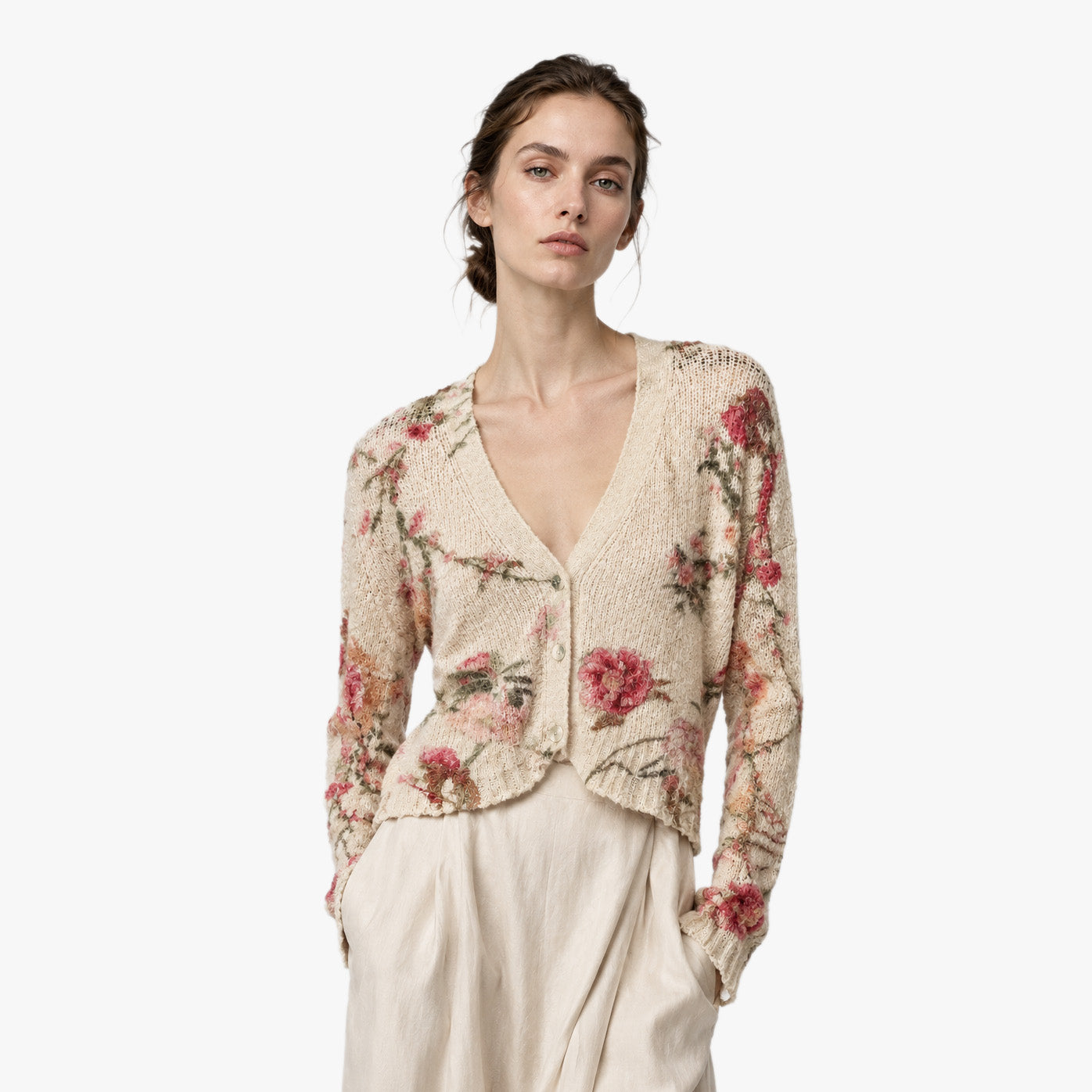 A woman modeling a cream-colored knit cardigan with a pink and red floral pattern, paired with light beige trousers, against a plain white background.