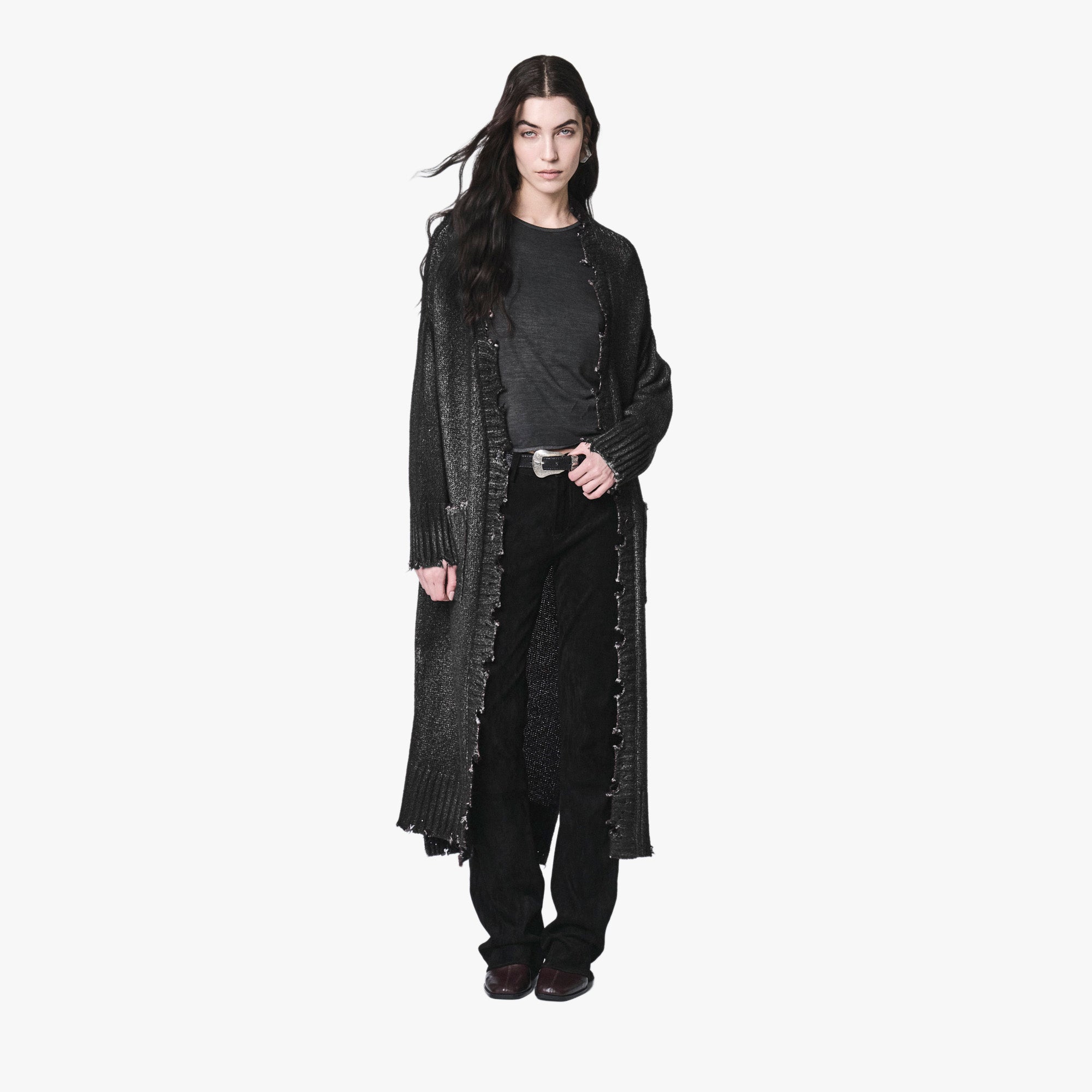 A model wearing a long, dark gray textured knit cardigan with fringe details over a black top and black pants, standing against a solid off-white background.