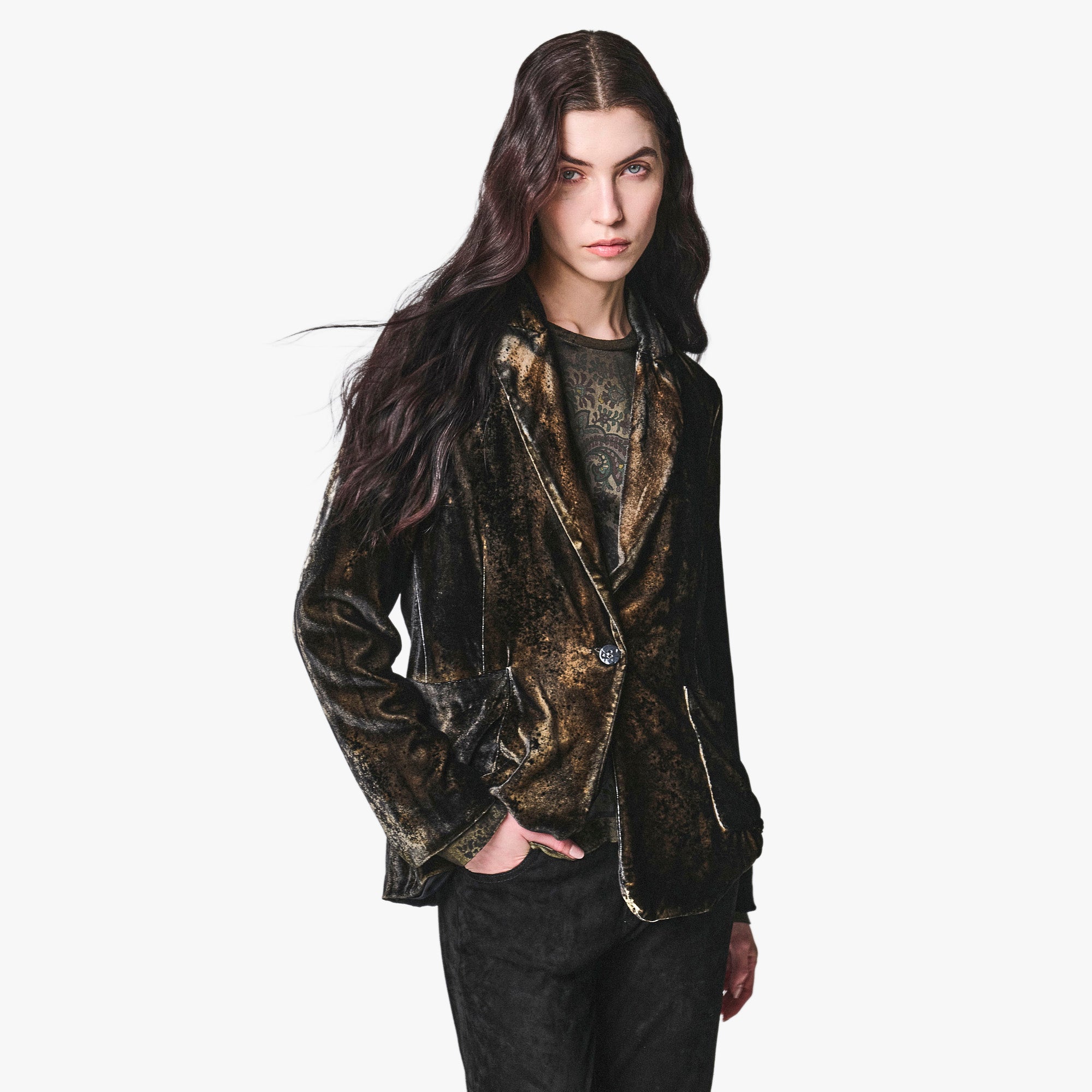 A model with long dark hair wearing a distressed, metallic gold and black velvet blazer over a dark graphic t-shirt and black pants, set against a plain white background.