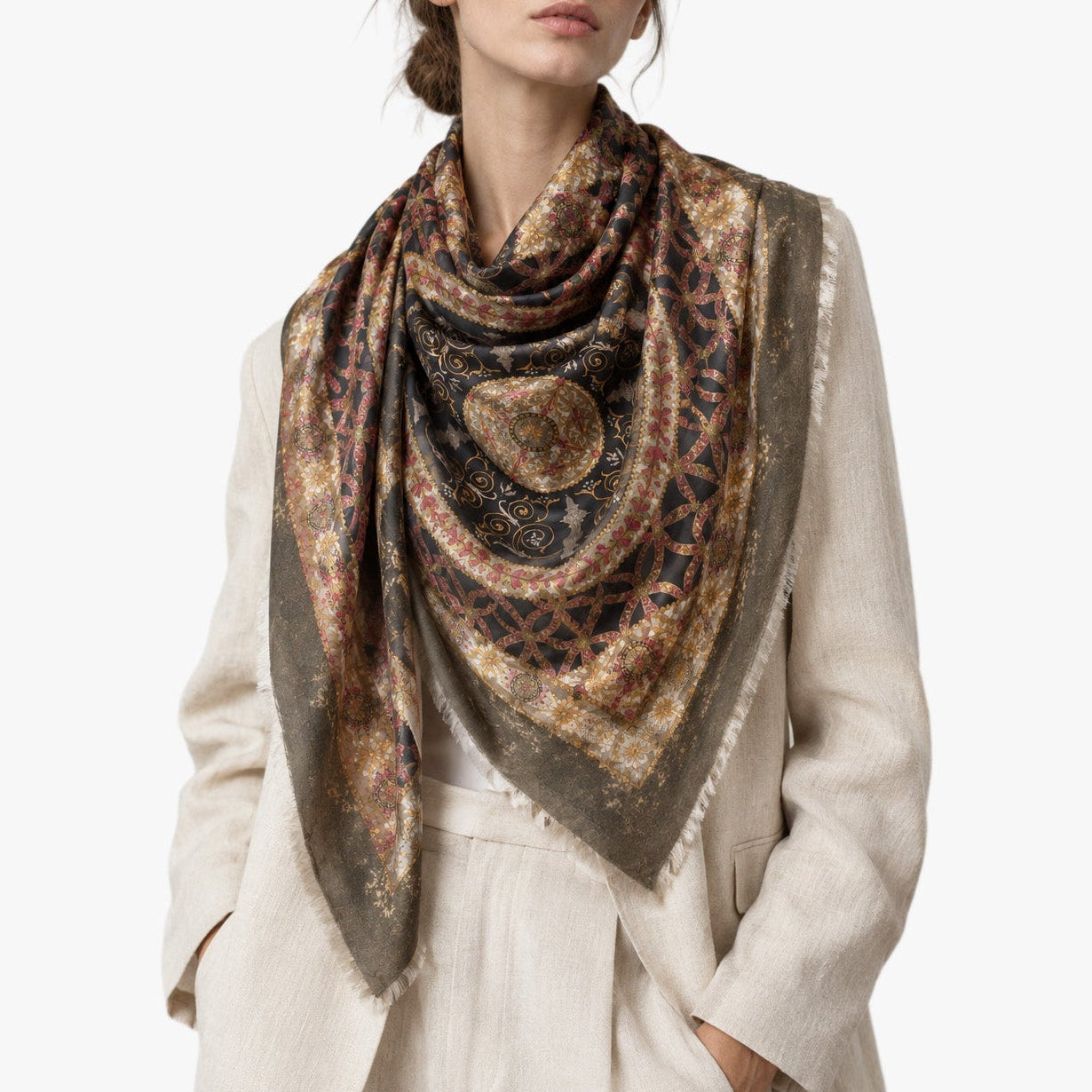 A woman wearing a cream-colored linen jacket and a large, patterned square scarf in earthy brown, gold, and navy tones, photographed against a solid white studio background.