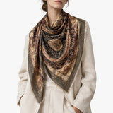 A woman wearing a cream-colored linen jacket and a large, patterned square scarf in earthy brown, gold, and navy tones, photographed against a solid white studio background.