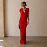 A woman modeling a vibrant red floor-length wrap dress with short sleeves and a draped, gathered skirt, standing in a minimalist beige-walled indoor setting.
