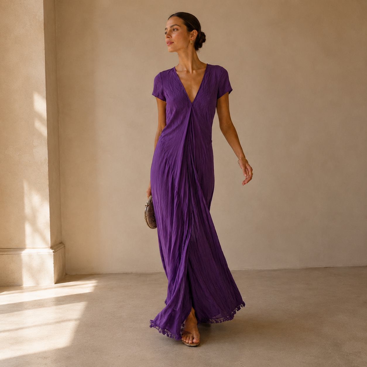 A woman models a purple crinkled maxi dress with short sleeves, a wrap-style V-neckline, and a side slit, standing in a minimalist beige room with soft window light.