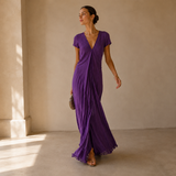 A woman models a purple crinkled maxi dress with short sleeves, a wrap-style V-neckline, and a side slit, standing in a minimalist beige room with soft window light.
