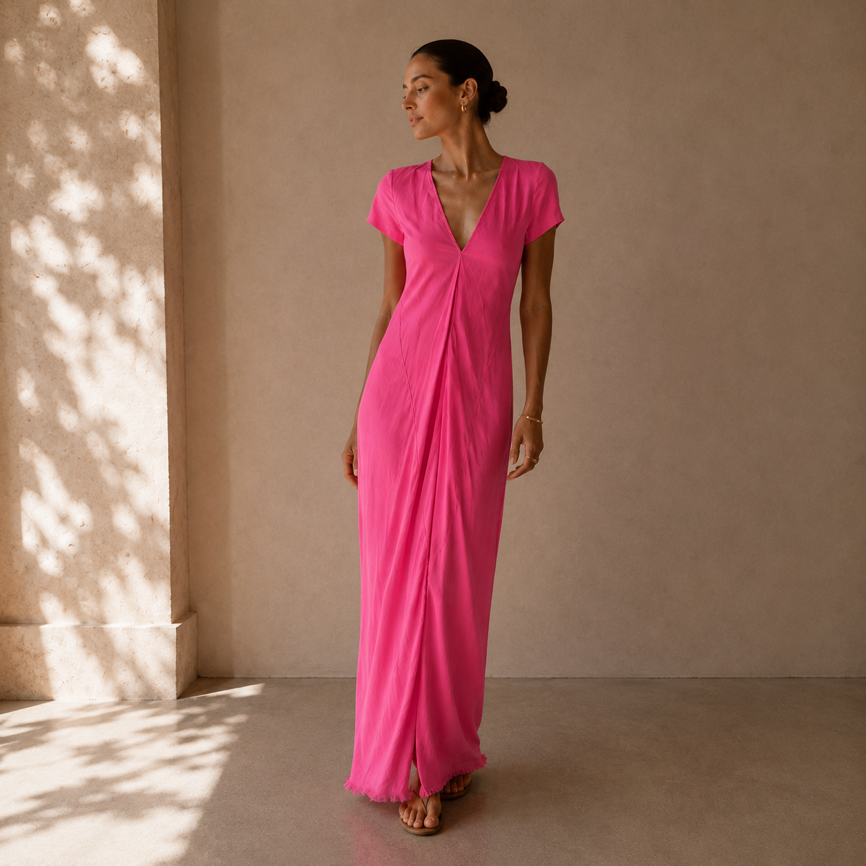 A woman wearing a bright pink short-sleeve maxi dress with a wrap-style front and V-neckline, standing in a sunlit beige interior space.