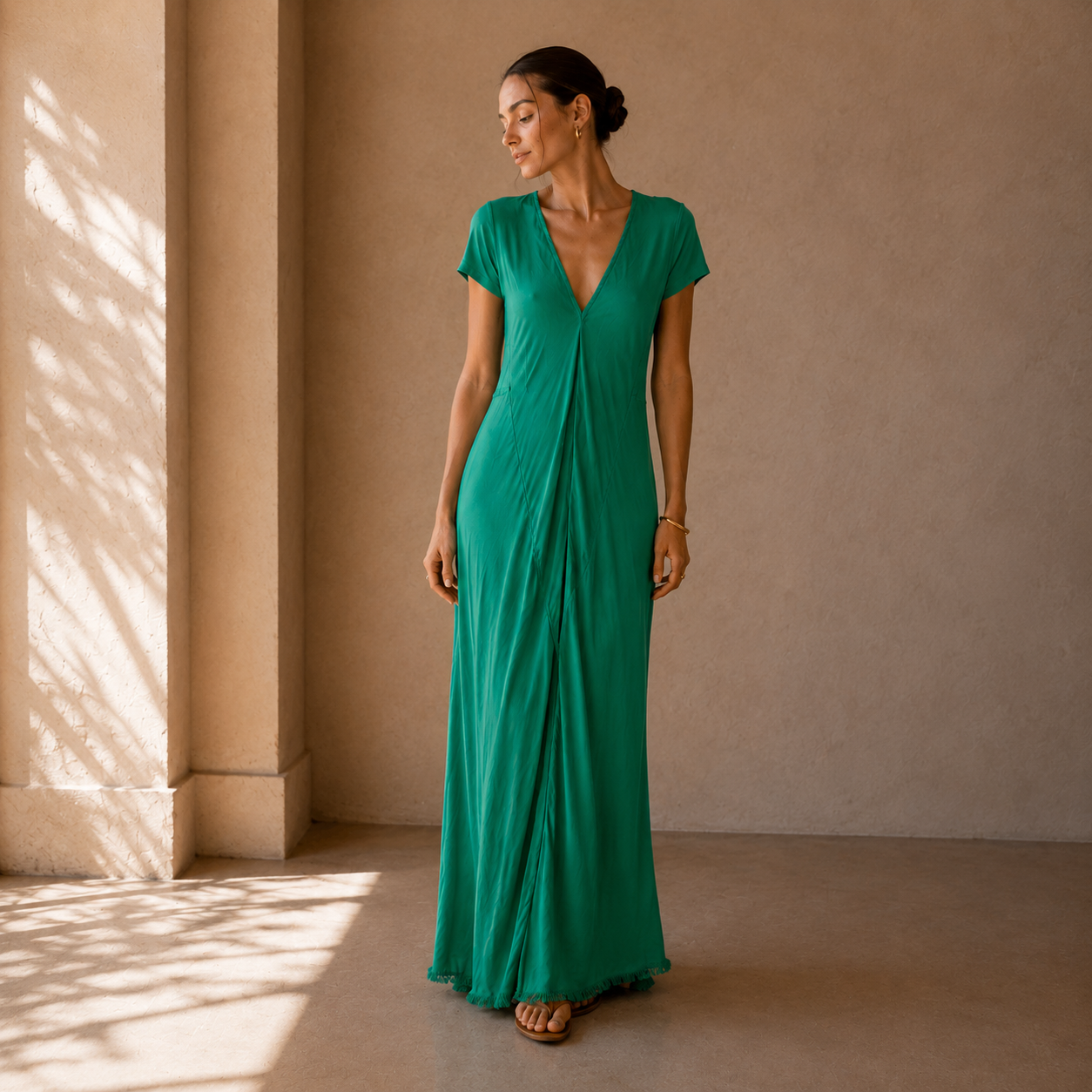 A woman wearing a flowing emerald green maxi dress with short cap sleeves and a deep V-neckline, standing in a sunlit architectural setting with a textured beige wall and floor.