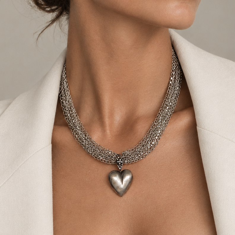 A close-up of a woman wearing a silver-toned multi-strand chain necklace with a polished heart-shaped pendant, styled with a white blazer against a neutral beige background.