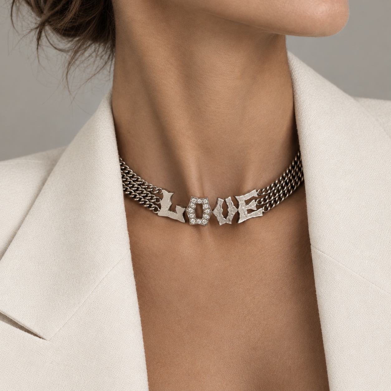 A close-up of a model wearing a silver-toned multi-strand choker necklace with the word 'LOVE' spelled out in raised letters, some embellished with small clear stones. The model is wearing a cream-colored blazer.