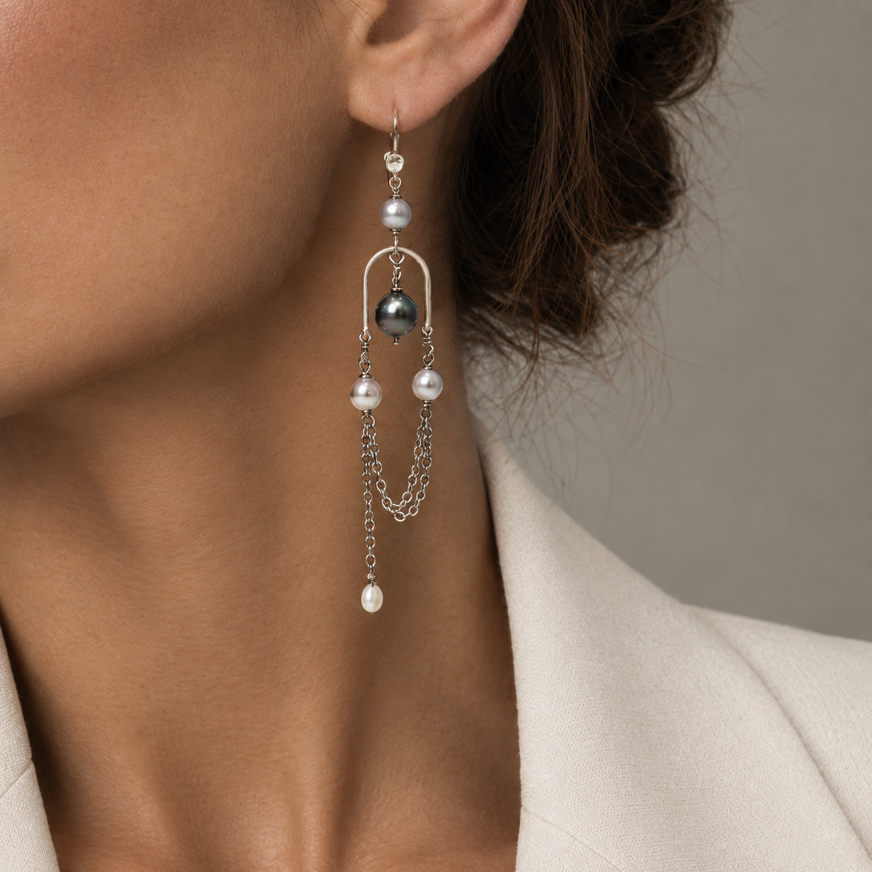 A close-up of a model wearing a long silver dangle earring with South Sea pearls and a Tahitian pearl, paired with a cream-colored blazer.