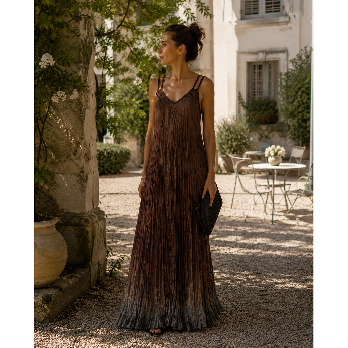A woman stands in a sunlit courtyard wearing a sleeveless, floor-length pleated dress in dark brown with a subtle gradient toward black at the hem. She is holding a small black clutch, with stone walls, greenery, and outdoor furniture in the background.
