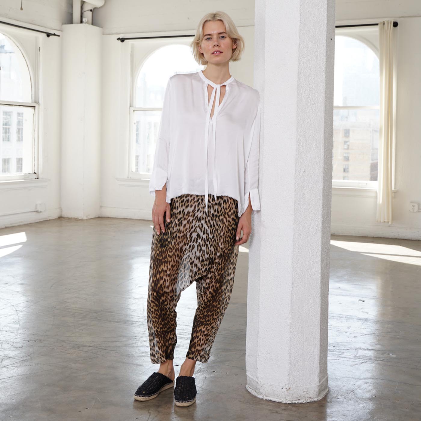 A woman stands in a bright, sunlit loft with white columns and large windows, wearing a loose white long-sleeve blouse with a tie neckline and drapey leopard-print harem pants with black slip-on shoes.