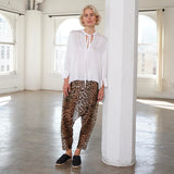 A woman stands in a bright, sunlit loft with white columns and large windows, wearing a loose white long-sleeve blouse with a tie neckline and drapey leopard-print harem pants with black slip-on shoes.