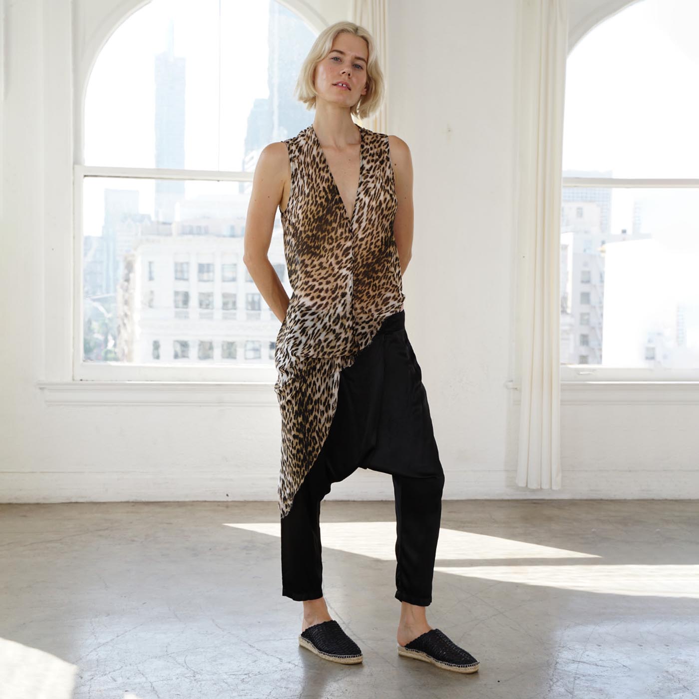 A woman stands in a bright, sunlit studio with large windows, wearing a sleeveless leopard-print top over a black raglan-style harem pant and black espadrille flats.