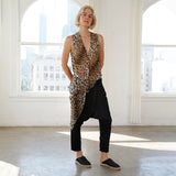 A woman stands in a bright, sunlit studio with large windows, wearing a sleeveless leopard-print top over a black raglan-style harem pant and black espadrille flats.