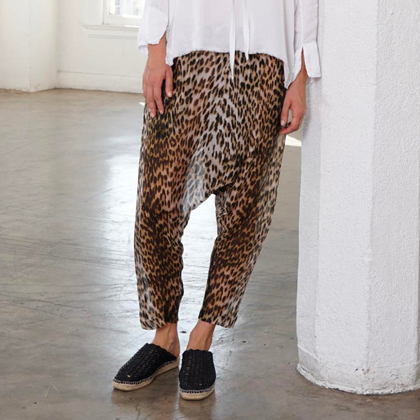 A woman stands in a bright, sunlit loft with white columns and large windows, wearing a loose white long-sleeve blouse with a tie neckline and drapey leopard-print harem pants with black slip-on shoes.