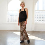 A woman stands in a bright white studio with large arched windows, wearing a black sleeveless top and leopard-print flared pants with black loafers.