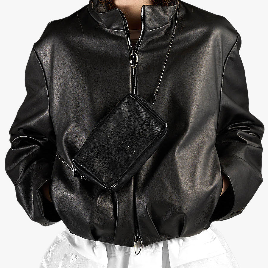 A person wearing a black faux-leather jacket with a front zipper and a matching black crossbody bag with a chain strap, shown against a plain light background.