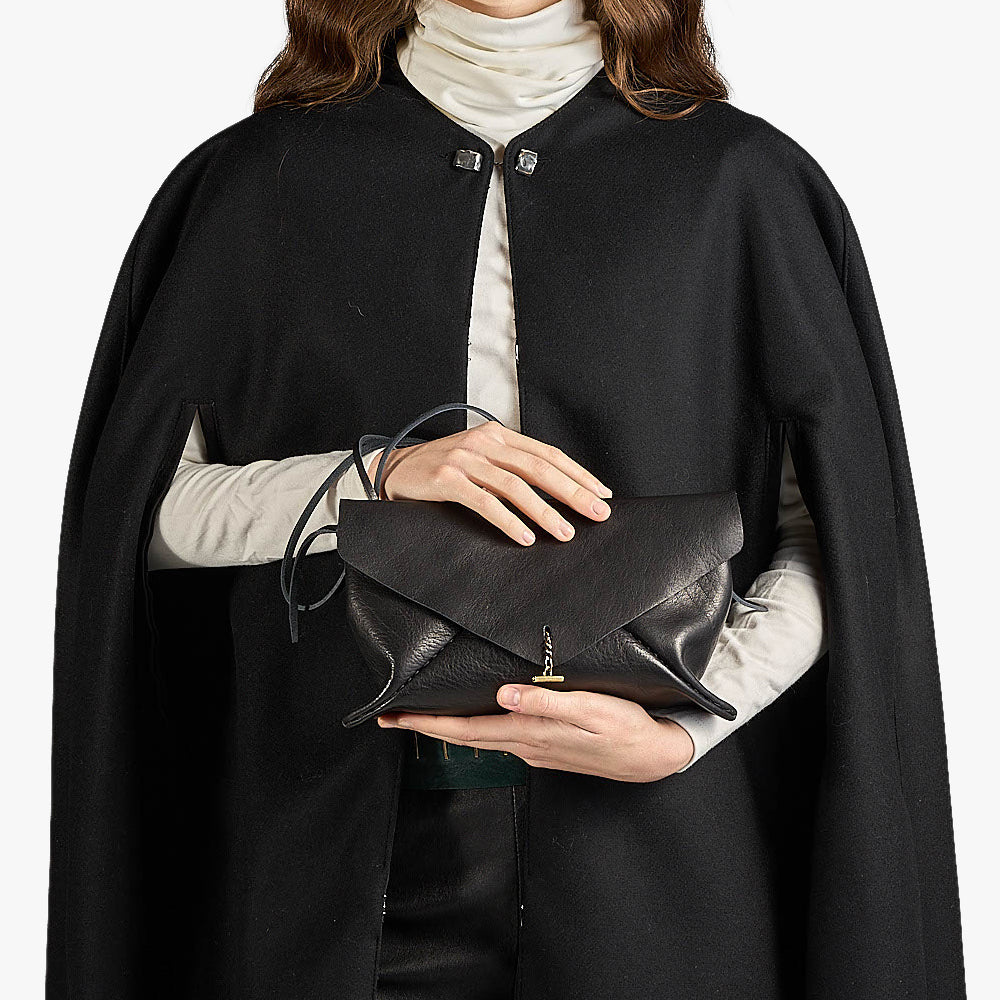 A model wearing a black cape-style coat over a cream turtleneck, holding a black leather envelope clutch with a gold-tone logo clasp.