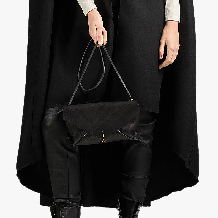 A person wearing a long black coat and black boots holds a black envelope-style shoulder bag with a thin strap against a plain light background.