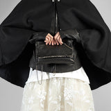 A person wearing a dramatic black cape-style jacket with wide sleeves and a white floral tulle skirt, holding a black leather crossbody bag with silver chain and zipper details against a plain gray studio background.