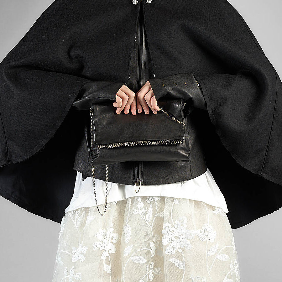 A person wearing a dramatic black cape-style jacket with wide sleeves and a white floral tulle skirt, holding a black leather crossbody bag with silver chain and zipper details against a plain gray studio background.
