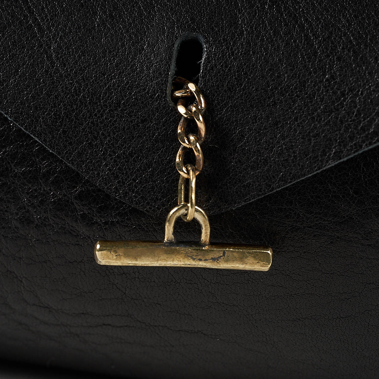 Close-up of a black textured leather bag featuring a gold-tone metal toggle clasp with a short chain link.