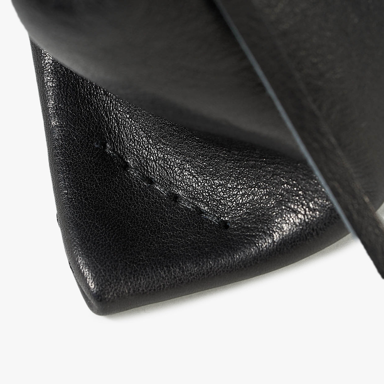 Close-up detail of a black textured leather product, showing a stitched corner and a dark strap against a clean white background.