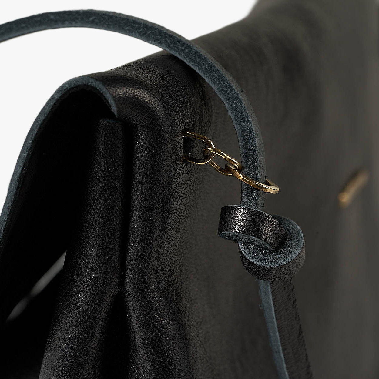 Close-up of a black leather shoulder bag showing the strap attachment with a gold-tone chain and ring, textured leather surface, and a white studio background.