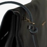 Close-up of a black leather shoulder bag showing the strap attachment with a gold-tone chain and ring, textured leather surface, and a white studio background.