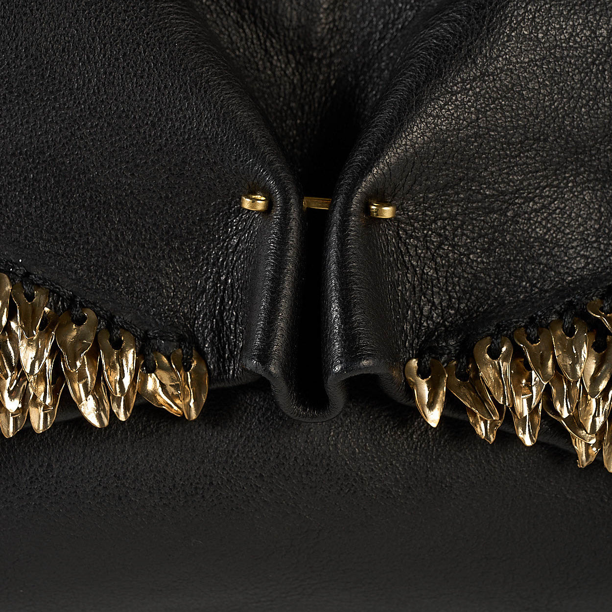 Close-up of a black textured leather clutch featuring a dramatic central fold and rows of sculptural gold-tone metallic shell embellishments along the lower edge.