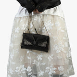A black leather clutch handbag with silver beaded wing details and a silver chain strap, held by a model wearing a sheer white floral tulle skirt and a black jacket.