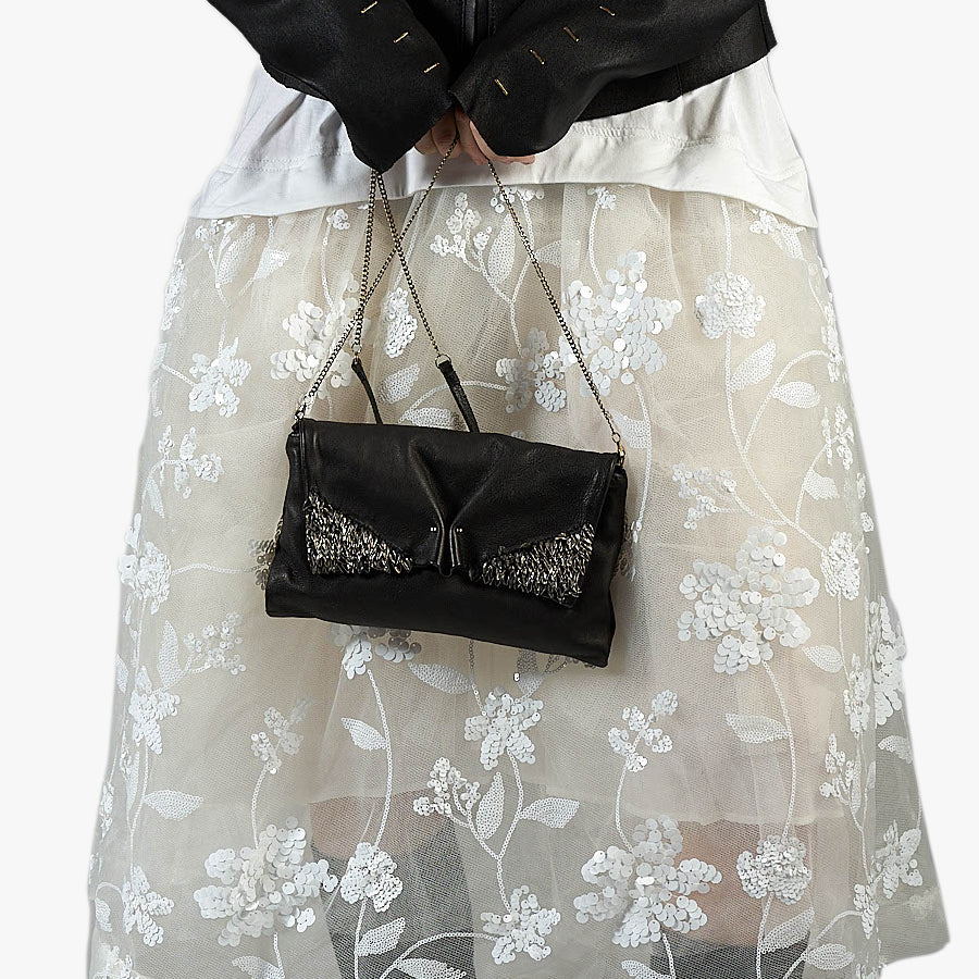 A black leather clutch handbag with silver beaded wing details and a silver chain strap, held by a model wearing a sheer white floral tulle skirt and a black jacket.