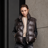 A model stands against a modern concrete and glass building wall, wearing a dark brown puffer vest over a cream sweater, and distressed denim pants.