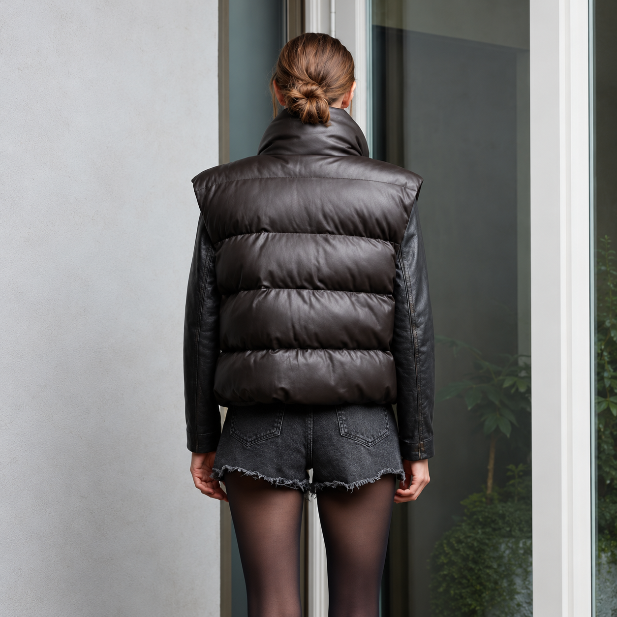 A model 's back facing the camera wearing a dark brown leather puffer vest with a high collar, worn over a black leather jacket.