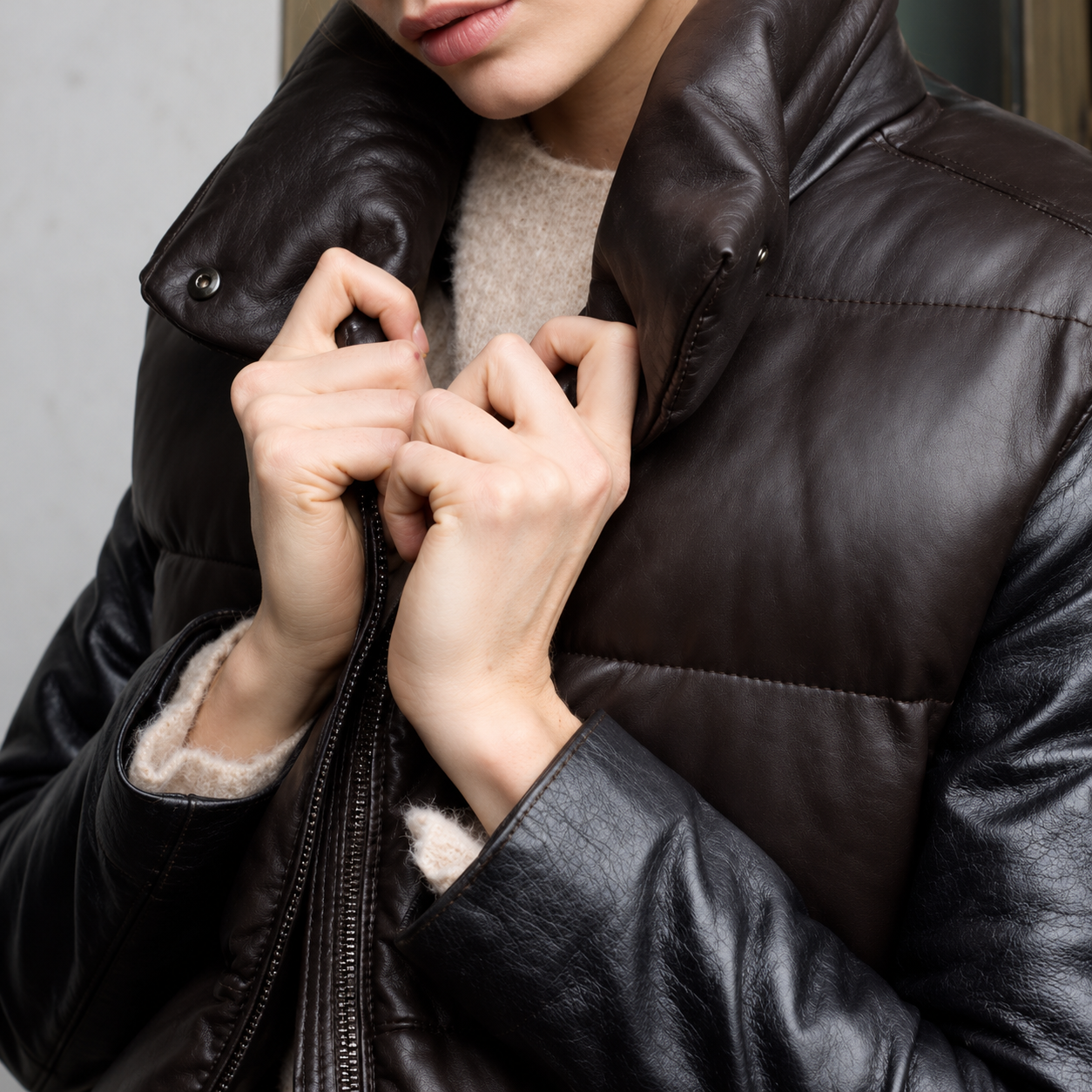 Close-up of a person wearing a dark brown leather puffer vest with a high collar and zipper, pulling the zipper with both hands.
The vest is worn over a beige knit sweater and black leather jacket.