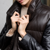 Close-up of a person wearing a dark brown leather puffer vest with a high collar and zipper, pulling the zipper with both hands.
The vest is worn over a beige knit sweater and black leather jacket.