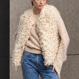 A woman stands outdoors against a concrete and black metal building wall, wearing a cream curly-texture shearling vest over a beige sweater, and blue jeans.