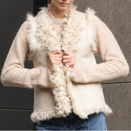 A woman stands outdoors in front of a modern stone and glass building, wearing a cream shearling and leather vest over a cream top with blue wide-leg jeans.