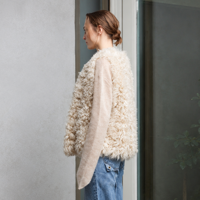 A woman stands in profile wearing a cream shearling vest over a light beige sweater, and blue jeans.