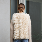 A woman stands with her back to the camera wearing a cream-colored shaggy shearling vest over a beige sweater and blue wide-leg jeans, positioned in front of a glass door on a concrete patio.