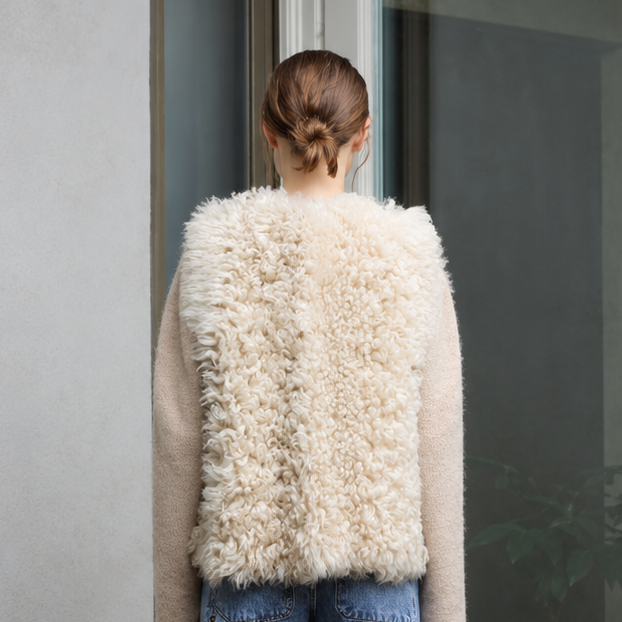A woman stands with her back to the camera wearing a cream-colored shaggy shearling vest over a beige sweater and blue wide-leg jeans, positioned in front of a glass door on a concrete patio.