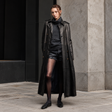 A model stands outdoors in front of a stone column wearing a long black leather trench coat over a dark charcoal turtleneck sweater, black shorts, sheer black tights, and black leather loafers.
