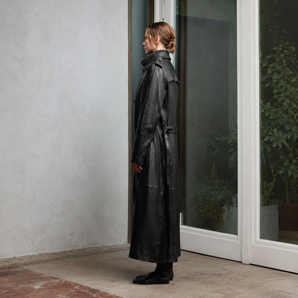 A person stands in profile wearing a long black leather trench coat, positioned beside a glass door in a modern interior setting with a textured light gray wall.