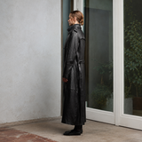 A person stands in profile wearing a long black leather trench coat, positioned beside a glass door in a modern interior setting with a textured light gray wall.
