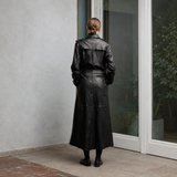 A woman viewed from behind wearing a long black leather trench coat, standing on a stone patio next to a glass door.