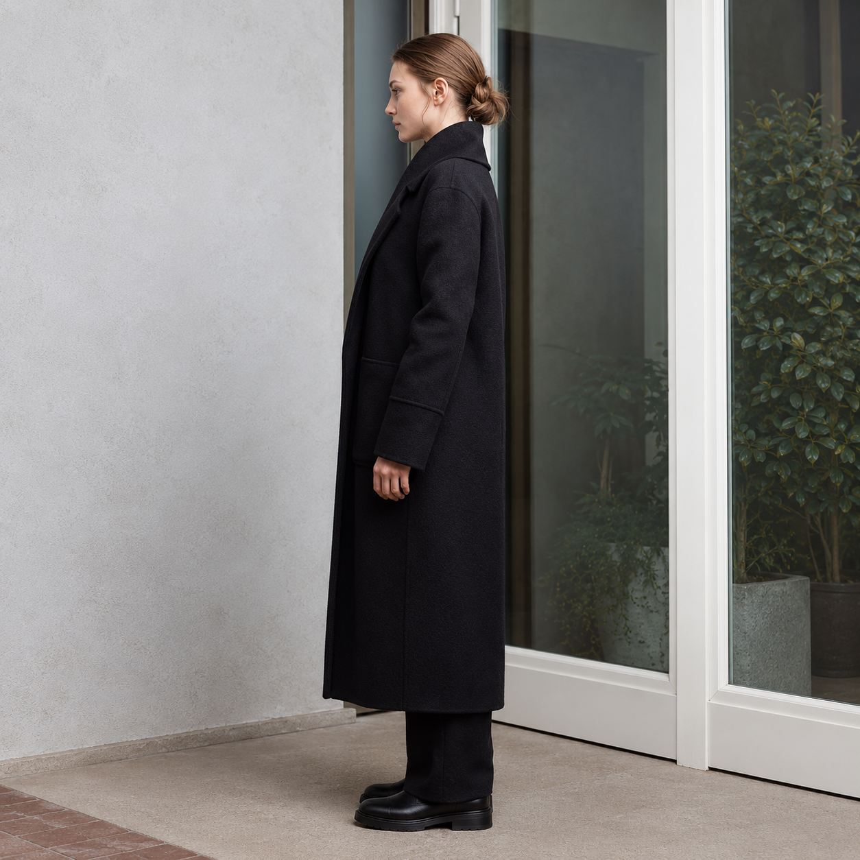 A woman stands in profile wearing a long black wool-blend coat, black trousers, and black shoes, positioned next to a glass door with a concrete wall on the left.