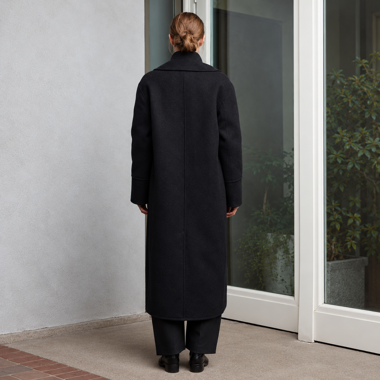 A woman viewed from behind wearing a long black coat and matching black trousers, standing on a tiled porch in front of glass doors.