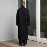 A woman viewed from behind wearing a long black coat and matching black trousers, standing on a tiled porch in front of glass doors.